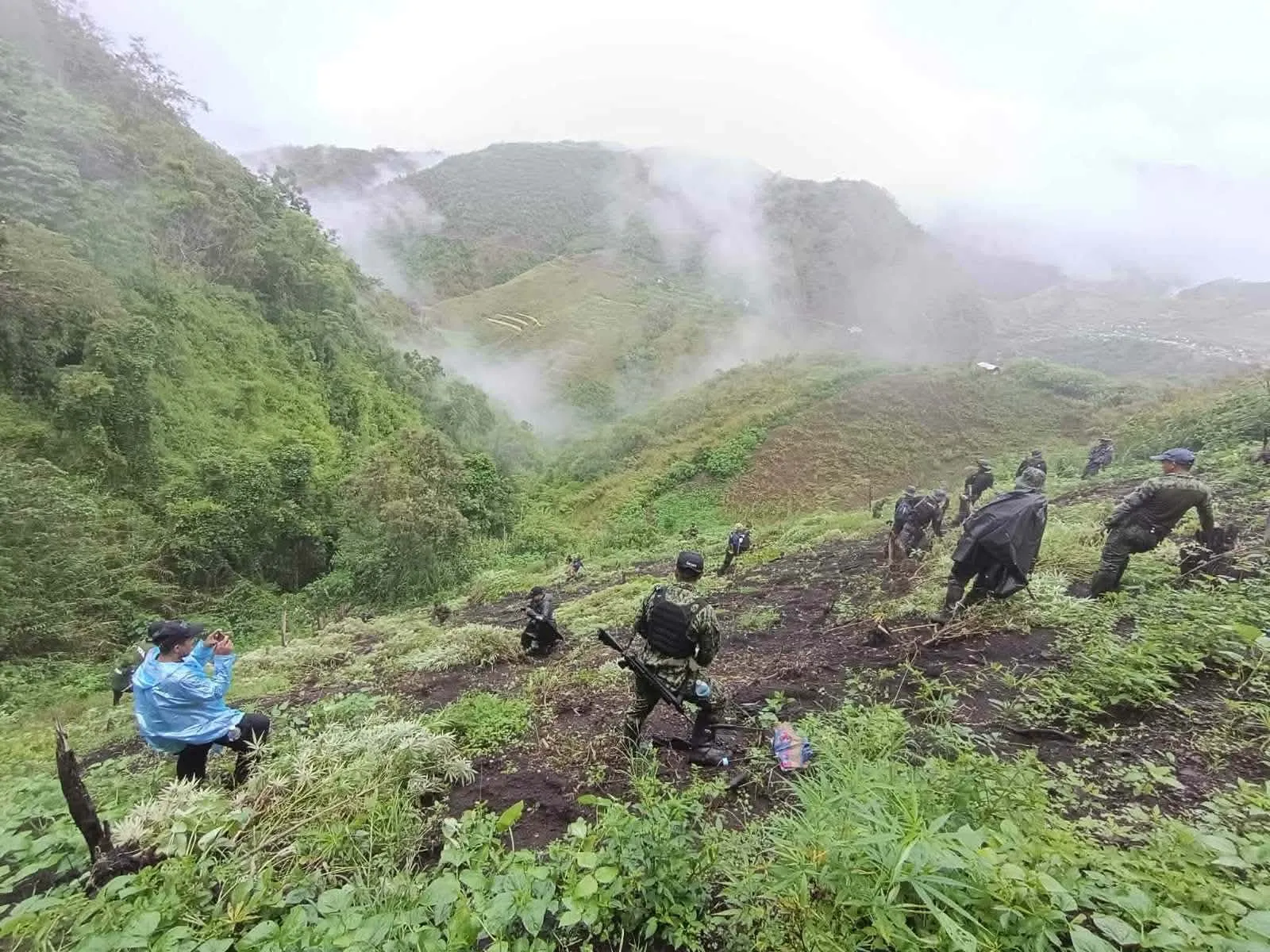 Police and drug agents in uniform climb a steep, cleared mountain slope in Tinglayan, with misty green ridges behind them